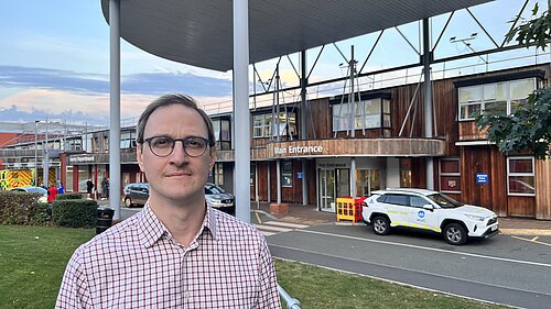 Ian Sollom standing across the road from the main entrance to Hinchingbrooke Hospital, 